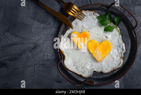 Uova fritte fatte in casa in una padella d'annata su sfondo grigio. Tuorlo a forma di cuore. Foto Stock