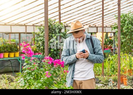 Giovane giardiniere maschio in un cappello che utilizza smartphone prende gli ordini e scatta foto di impianti per l'elaborazione degli ordini dei clienti in serra. Vendita di orn Foto Stock