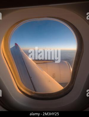 Vista attraverso una finestra di un aereo, cielo blu, nuvole sullo sfondo. Sole serale che splende su un'ala di aerei, scenario maestoso all'orizzonte. Foto Stock