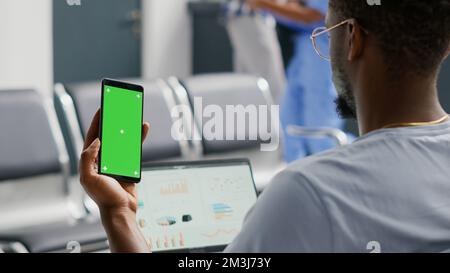 Paziente maschio che utilizza un modello con schermo verde sullo smartphone mentre attende nell'area di attesa della struttura. Giovane uomo che guarda il display mockup isolato e la chiave chroma, spazio vuoto del copyspace sullo schermo del telefono cellulare. Foto Stock