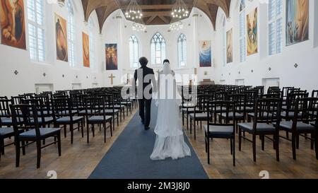 Cerimonia di nozze solo per sposa e sposo in cappella. Azione. Vista posteriore di un uomo in tuta e di una donna in vestito bianco che camminano insieme lungo le righe vuote i Foto Stock