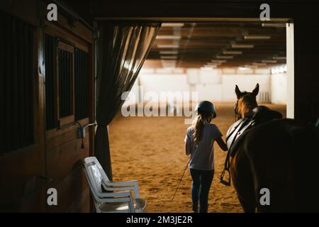 La ragazza conduce il cavallo con la sella nell'arena di equitazione con il casco inserito Foto Stock