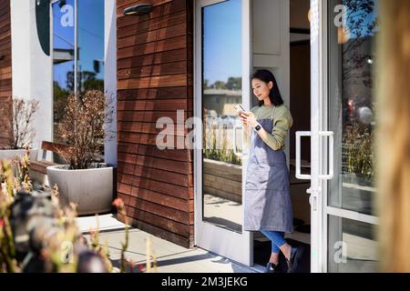 Sorridente proprietario del negozio di caffè che usa il telefono cellulare mentre si appoggia alla porta Foto Stock