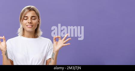 Tranquilla bionda femminile dall'aspetto gradevole con occhi di chiusura della maglietta bianca sorridente alleviato e felice sensazione soddisfatto con la vita in piedi posa del loto Foto Stock