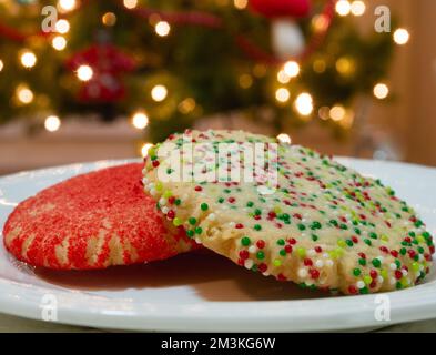 Primo piano estremo di due biscotti di zucchero di Natale decorati con zucchero rosso e spruzzi rossi, bianchi e verdi con le luci di un albero di Natale in th Foto Stock