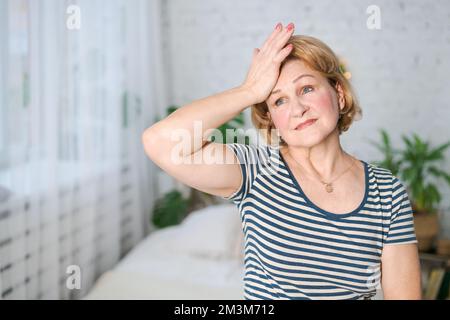 Donna matura infelice che tocca la testa mentre sente mal di testa grave mentre si siede da sola a casa, ha sconvolto le donne anziane che soffrono di emicrania o di pressione alta, concetto di problema di salute Foto Stock