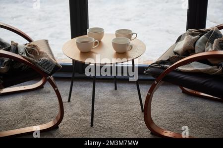 Quattro tazze di tè incompiuto su un piccolo tavolo e sedie dal design accogliente con plaid a scacchi nel caffè. Vista frontale di tazze bianche su tavolo di legno con sedie di fronte alla finestra durante il giorno. Concetto di riposo. Foto Stock