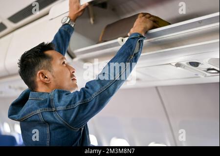 Bel passeggero asiatico maschio mettere il suo bagaglio a mano sul armadietto superiore prima di decollare. concetto di volo, aereo, trasporto Foto Stock