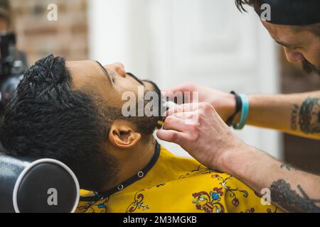 Barbiere professionale con tatuaggi rasa la barba del cliente indiano con pettine e rifinitore. Foto di alta qualità Foto Stock