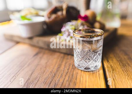 Stinco di maiale arrosto servito in modo moderno su un piatto di legno sfocato sullo sfondo. Bicchiere con acqua o vodka in primo piano. Foto di alta qualità Foto Stock