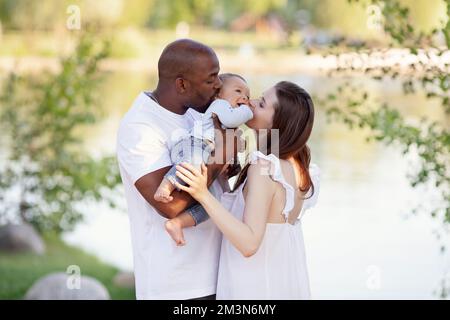 Felice famiglia multietnica. Madre caucasica e Padre afroamericano baciano il bambino piccolo. Genitori, Ritratto di mamma, papà e bambino sulle mani sopra la natura b Foto Stock