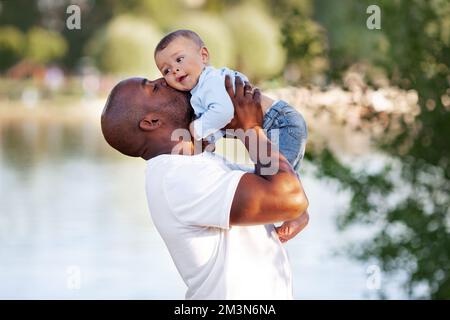 Il padre afroamericano bacia, tiene le braccia e ama il figlio bambino gara mista all'aperto in estate Foto Stock