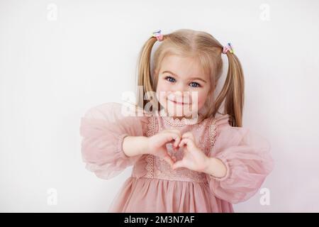 La bambina in abito rosa con coda di cavallo mostra il segno del cuore con le mani isolate su sfondo bianco Foto Stock