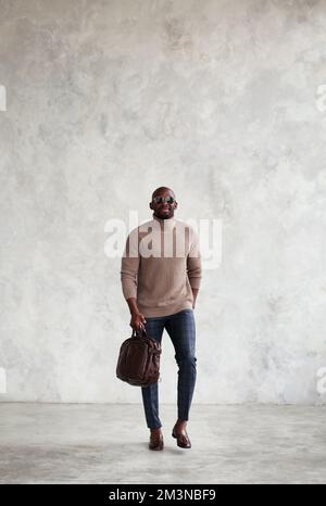 Uomo alla moda che cammina contro le pareti grigie, indossa abiti eleganti, borsa in pelle marrone e loafer, pullover dolcevita. Uomo stile di moda Foto Stock