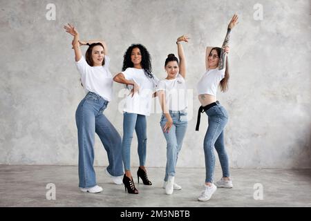 Donne multietniche sorridenti posano, ballano, alzano le mani sullo sfondo grigio dello studio. Il gruppo di ragazze è simile vestito in jeans e T-shirt bianca Foto Stock