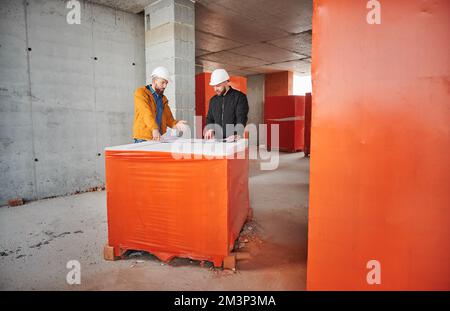 Uomo che discute il progetto architettonico di nuovo appartamento con il lavoratore di costruzione, in piedi all'interno dell'edificio di appartamento in costruzione e parlare con il costruttore. Foto Stock