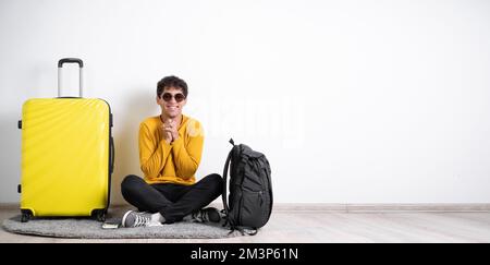 Il giovane viaggiatore con una valigia soddisfatto di un'espressione positiva si siede isolato su uno sfondo bianco tiene le mani vicino al petto Foto Stock