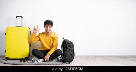 Mano del viaggiatore che mostra il segno del rock n roll o che dà il gesto delle corna del diavolo. Giovane viaggiatore seduto con valigia su sfondo isolato bianco letto Foto Stock