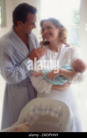 Coppia più anziana in abbigliamento da sonno tenendo un neonato di fronte alla finestra con un look morbido. La donna sorride all'uomo Foto Stock