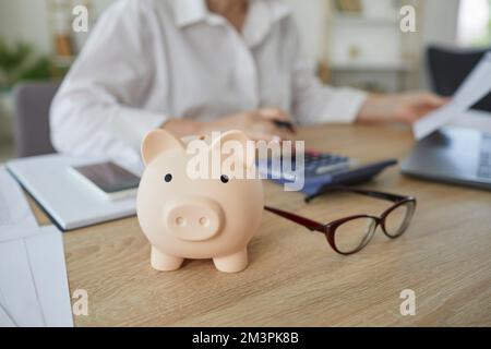 Piggy Bank in forma di maiale sulla scrivania di finanziere femmina che lavora con la calcolatrice. Foto Stock