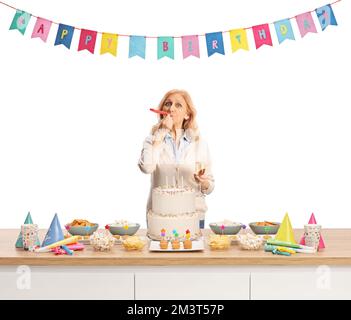 Casual donna matura soffiare un compleanno corno festa dietro un bancone con torta isolato su sfondo bianco Foto Stock