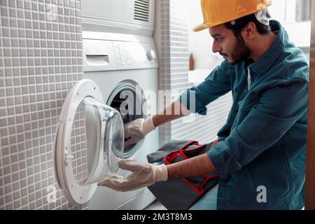 Il riparatore messo a fuoco in vestito di lavoratore sta fissando la lavatrice nella stanza da bagno Foto Stock