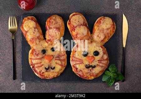 Deliziosa pizza al coniglietto con salsa di pomodoro, salame, formaggio, olio d'oliva, olive nere e basilico su piatto d'ardesia. Cibo idea per Capodanno Foto Stock