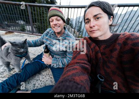 Giovane donna caucasica 30s si siede con suo marito e gatto godendosi una bevanda mentre ha il suo gatto grigio blu russo sul guinzaglio nella foresta selvaggia mentre a. Foto Stock