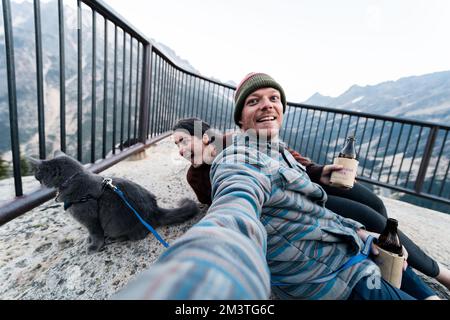 Giovane donna caucasica 30s si siede con suo marito e gatto godendosi una bevanda mentre ha il suo gatto grigio blu russo sul guinzaglio nella foresta selvaggia mentre a. Foto Stock