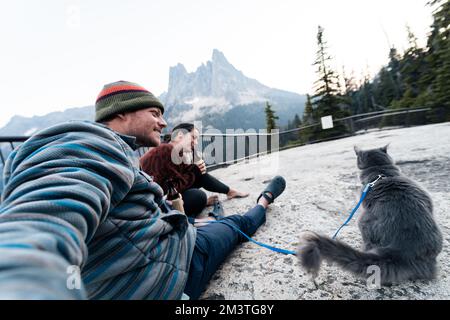 Giovane donna caucasica 30s si siede con suo marito e gatto godendosi una bevanda mentre ha il suo gatto grigio blu russo sul guinzaglio nella foresta selvaggia mentre a. Foto Stock