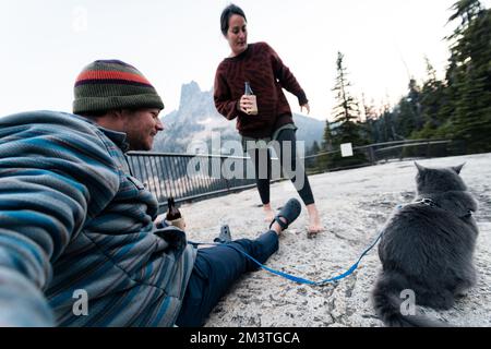 Giovane donna caucasica 30s si siede con suo marito e gatto godendosi una bevanda mentre ha il suo gatto grigio blu russo sul guinzaglio nella foresta selvaggia mentre a. Foto Stock