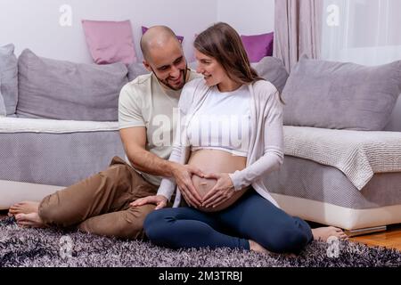 Una giovane coppia felice seduta sul pavimento, che si prepara al parto. Il marito amorevole carezza dolcemente il ventre incinta di sua moglie. Foto Stock
