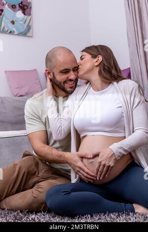 Una giovane coppia felice seduta sul pavimento, che si prepara al parto. Il marito amorevole carezza dolcemente il ventre incinta di sua moglie. Foto Stock