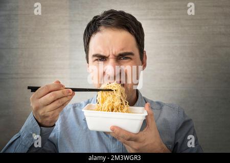 Giovane uomo che mangia spaghetti istantanei mentre lavora in ufficio. Pranzo in ufficio. cibo spazzatura insapore Foto Stock