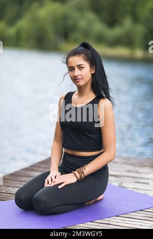 Ritratto di una giovane bella allenatrice di yoga femminile seduta su una passerella di legno sulla riva di un lago Foto Stock