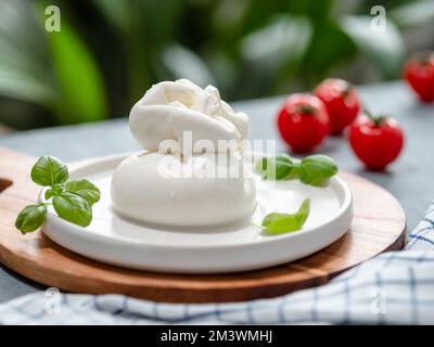 Formaggio italiano fresco su piatto circolare bianco. Pallina bianca di burrata o burrata, basilico decorato e pomodori. Pallina di burrata bianca morbida estetica su sfondo verde sfocato, spazio copia Foto Stock