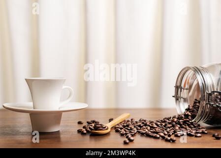 Tazza di caffè accanto a un vaso capovolto con chicchi di caffè tostati, alcuni su un cucchiaio di legno. Foto Stock