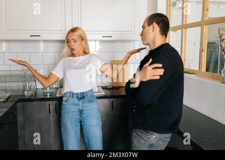 Stanco frustrato marito caucasico ignorando arrabbiato bionda moglie che sostiene incolpare l'uomo sconvolto di problemi in piedi in cucina. Foto Stock