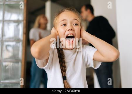 Primo piano ritratto di eccitato rovesciato figlia piccola urlare guardando lontano, coprendo le orecchie mentre genitori arrabbiati litigare e combattere sullo sfondo a. Foto Stock