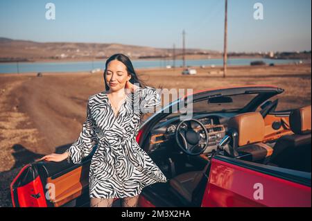 Felice bruna sorridente donna conducente in abito di moda in posa con la nuova cabriolet rossa sulla costa della spiaggia, godendo e pronto per il viaggio. Corsi di guida e concetto di assicurazione sulla vita. Stile di vita glamour Foto Stock