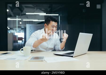 Un giovane asiatico si siede in ufficio al tavolo, durante le ore lavorative guarda una partita di calcio, gare sportive, fa scommesse, alliera e gioisce sul suo computer portatile. Foto Stock