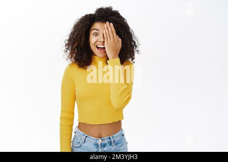 Donna afroamericana sorridente con capelli ricci, pelle chiara e luminosa, coprire, nascondere metà del viso, un occhio con palmo, guardare felice la macchina fotografica, in piedi Foto Stock