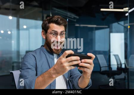 Uomo d'affari maturo con la barba sta giocando a gioco su smartphone, l'uomo si diverte durante la pausa sul posto di lavoro, uomo d'affari all'interno di un ufficio moderno. Foto Stock