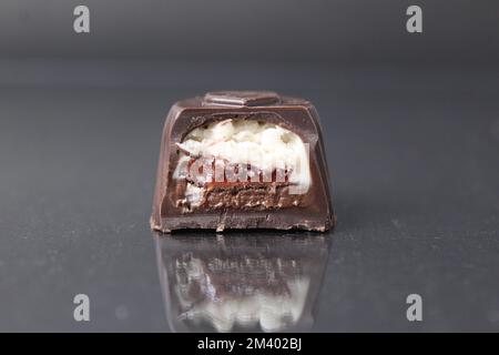 una fetta cremosa di caramelle al cioccolato in una sezione su sfondo nero e una caramella intera l'una accanto all'altra. Ripieno di cioccolato bianco al latte con cocco Foto Stock