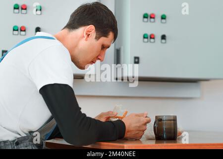Pausa pranzo. Mangiare sul posto di lavoro durante il lavoro. L'uomo caucasico in tuta da lavoro siede al tavolo nella sala di produzione e mangia dal contenitore. Il lavoratore ha pranzo in sala pausa. Foto Stock