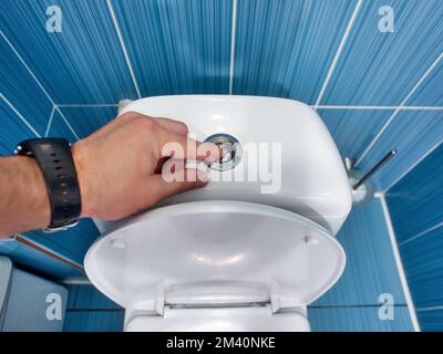 L'uomo preme il pulsante del gabinetto con la mano e scarica l'acqua nel gabinetto. Vista in prima persona. Spazio interno del bagno per gli uomini.. Foto Stock