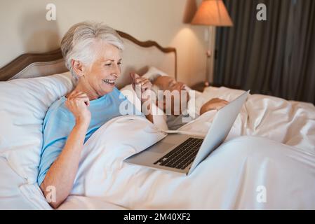 OH, non vedo l'ora di mostrargli questo. una donna anziana seduta a letto e che usa un computer portatile mentre suo marito dorme accanto a lei. Foto Stock