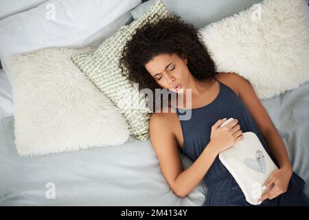 OW il mio pancino fa male. una giovane donna attraente che soffre di crampi d'epoca mentre sdraiata a letto con una bottiglia d'acqua calda. Foto Stock