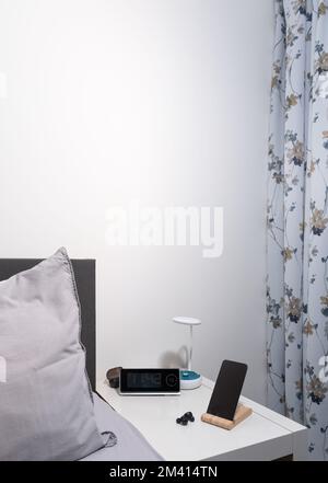 smartphone sul comodino con lampada, cuffia, orologio e letto come sfondo. verticale Foto Stock