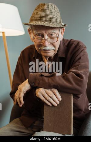 Uomo anziano seduto da solo sulla poltrona nel soggiorno indossando occhiali e tenendo un libro, guardando attentamente la macchina fotografica Foto Stock
